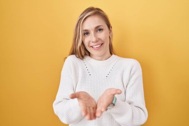 Sarı arka planda beyaz süveter giyen, avuçlarını birbirine değdirip el hareketi yapan beyaz bir kadın. Bekleme ve koruma 