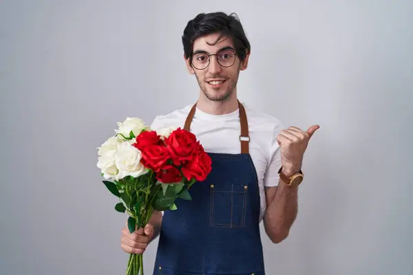 Young hispanic man holding bouquet of white and red roses pointing to the back behind with hand and thumbs up, smiling confident