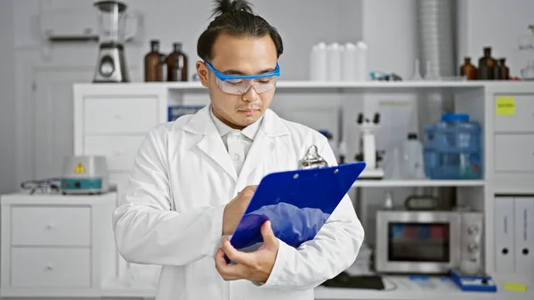 Serious young chinese man scientist, with a fashionable pigtail, diligently taking notes on a checklist in the meticulous world of lab research