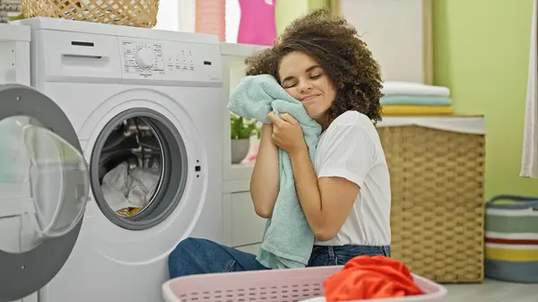 Young beautiful hispanic woman washing clothes touching face with clean towel at laundry room
