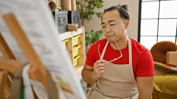 Serious young chinese man artist, with a handsome pigtail hairstyle, sitting relaxed yet concentrated, looking at his drawing in the art academy studio, deep in thought and doubt.