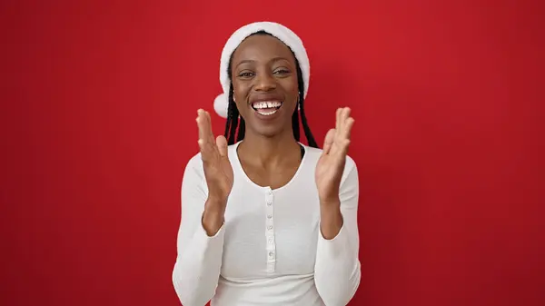 クリスマスの帽子をかぶったアフリカ系アメリカ人女性は 孤立した赤い背景の上に手をぶつける — ストック写真