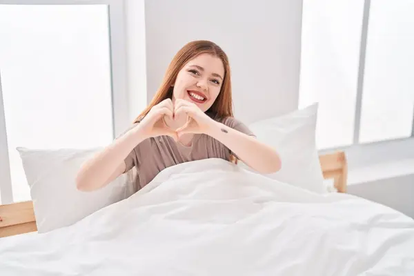 Young redhead woman doing heart gesture sitting on bed at bedroom
