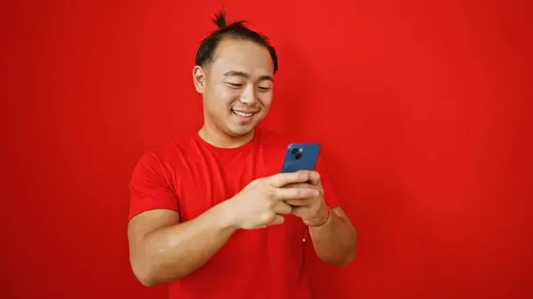 Cheerful young asian guy joyfully texting on his phone, isolated against a vibrant red background