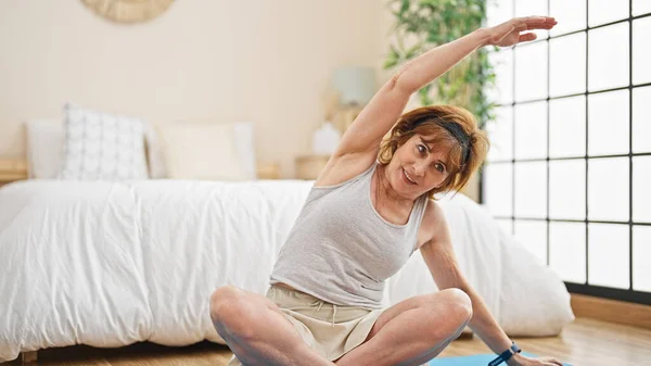Middle age woman stretching back sitting on yoga mat at bedroom