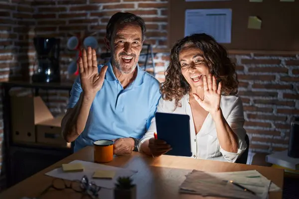 Middle age hispanic couple using touchpad sitting on the table at night waiving saying hello happy and smiling, friendly welcome gesture