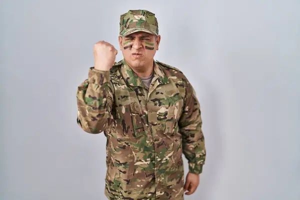 Hispanic young man wearing camouflage army uniform angry and mad raising fist frustrated and furious while shouting with anger. rage and aggressive concept.