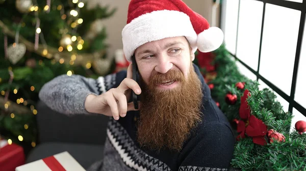 Young redhead man talking on smartphone sitting on sofa by christmas tree at home