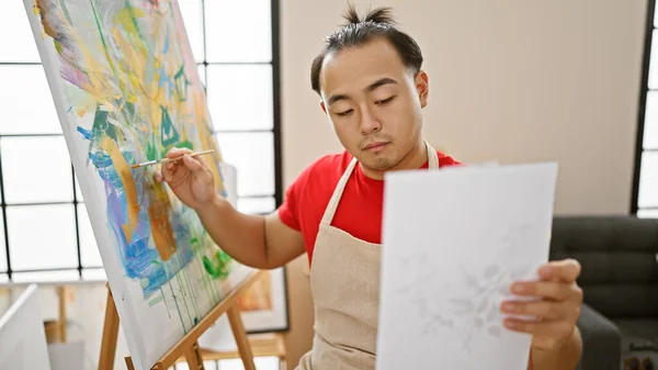 Young, handsome chinese artist man seriously concentrating, looking at drawing on paper in indoor art studio, enveloped in creativity amid learning