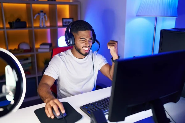 Young latin man streamer playing video game with winner expression at gaming room