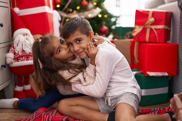 Brother and sister kissing and hugging each other sitting on floor by christmas gifts at home