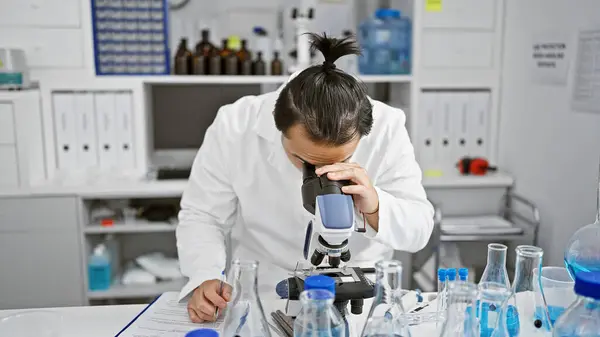 Serious young chinese man, a professional scientist, concentrating at a microscope in a bustling lab - a marriage of medicine and technology
