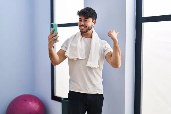 Hispanic man with beard doing video call with smartphone at the gym pointing thumb up to the side smiling happy with open mouth
