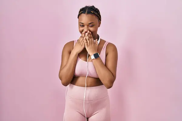 African american woman with braids wearing sportswear and headphones laughing and embarrassed giggle covering mouth with hands, gossip and scandal concept