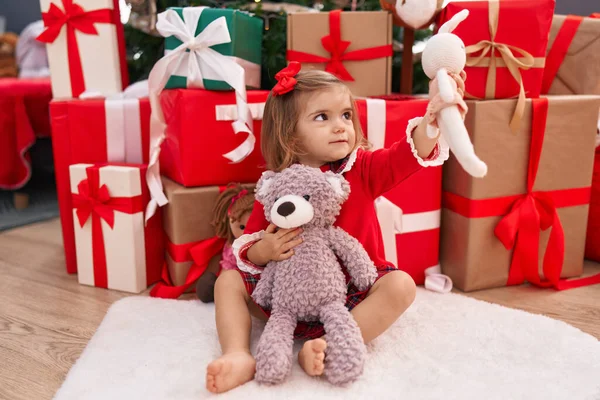 Sevimli sarışın bebek elinde oyuncak ayıyla evde Noel hediyeleriyle oturuyor.