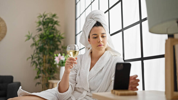 Молодая красивая латиноамериканка в халате пьет шампанское, используя смартфон в спальне.