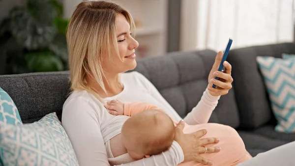 Mãe Filha Sentadas Sofá Amamentando Bebê Usando Smartphone Casa — Fotografia de Stock