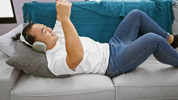 stock image Joyful young chinese man lying on the sofa, confidently listening and dancing to music at home, immersed in the audio world with headphones, expressing positive lifestyle and happiness.