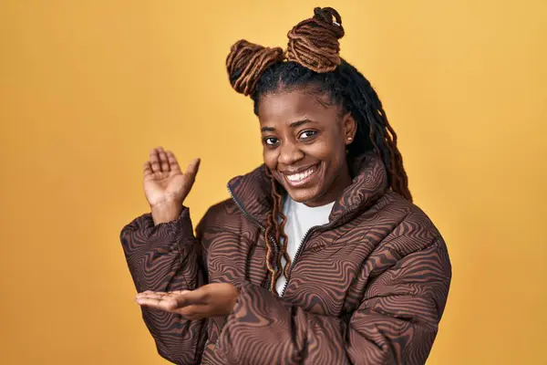 オープンハンドで笑顔の自然に入るために招待黄色の背景の上に立っている編組の髪を持つアフリカの女性 — ストック写真