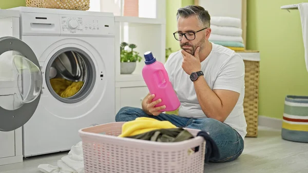 Grey-haired man washing clothes holding detergent bottle thinking at laundry room