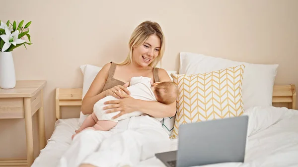 Mother and daughter sitting on bed breastfeeding baby having video call at bedroom