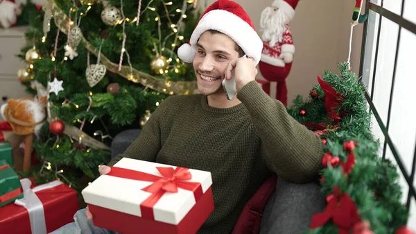Νεαρός Ισπανόφωνος Μιλάει Στο Smartphone Κάθεται Στον Καναπέ Κρατώντας Χριστουγεννιάτικο — Φωτογραφία Αρχείου