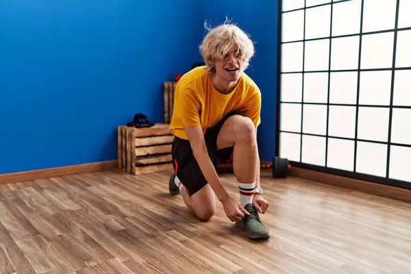 Young blond man smiling confident tying shoe at sport center