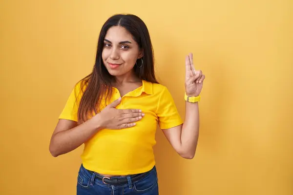 年轻的阿拉伯女人站在黄色的背景上微笑着 双手托着胸膛和手指发誓 宣誓效忠 — 图库照片
