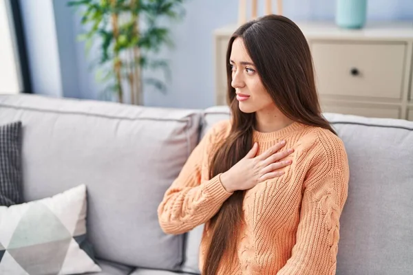 Young beautiful hispanic woman sitting on sofa suffering heart attack at home