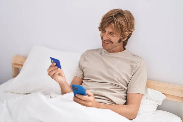 Genç Adam Yatakta Oturan Akıllı Telefon Kredi Kartı Kullanıyor — Stok fotoğraf