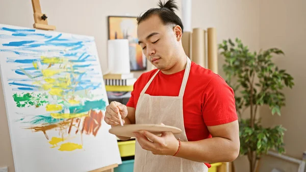 Serious-faced young chinese artist, clutching paintbrush and palette, commands the studio space with a relaxed stance