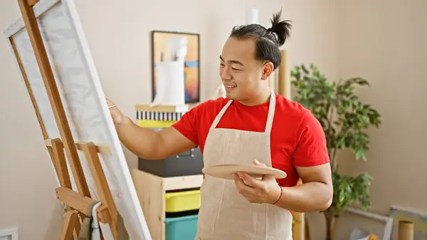 Confident young chinese artist smiling as he enjoys drawing in a vibrant art studio, embodying creativity with a brush and canvas