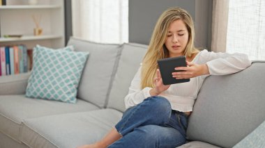Young blonde woman using touchpad sitting on sofa at home