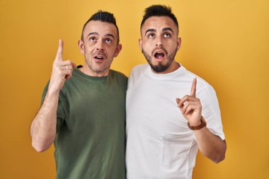 Sarı arka planda duran eşcinsel çift şaşırmış ve şaşırmış bir şekilde yukarı bakıp parmaklarını göstererek kollarını kaldırmış.. 
