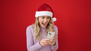 Yılbaşı şapkası takan genç sarışın kadın, izole edilmiş kırmızı arka planda sürpriz bir ifadeyle akıllı telefon kullanıyor.