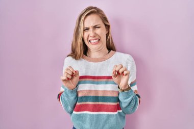 Young blonde woman standing over pink background disgusted expression, displeased and fearful doing disgust face because aversion reaction. 