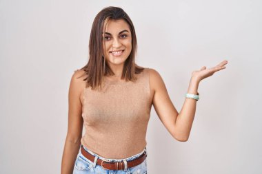 Young hispanic woman standing over white background smiling cheerful presenting and pointing with palm of hand looking at the camera. 