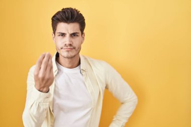 Young hispanic man standing over yellow background doing italian gesture with hand and fingers confident expression 