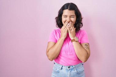 Young hispanic woman standing over pink background laughing and embarrassed giggle covering mouth with hands, gossip and scandal concept 