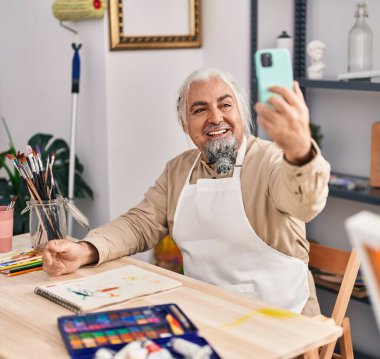 Orta yaşlı, gri saçlı, gülümseyen bir sanatçı sanat stüdyosundaki akıllı telefondan özçekim yapıyor.