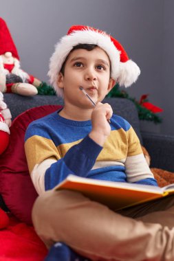 Evdeki Noel süslemeleriyle birlikte kanepeye oturan sevimli İspanyol çocuk deftere yazı yazıyor.