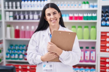 Young beautiful hispanic woman pharmacist smiling confident holding clipboard at pharmacy