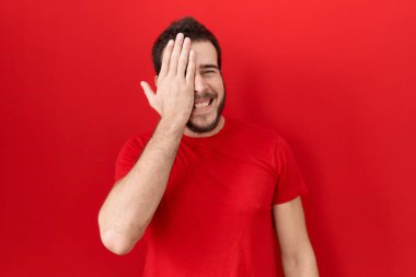 Latin asıllı genç bir adam gündelik kırmızı tişört giyiyor. Bir gözünü eliyle kapatıyor. Yüzünde kendine güvenen bir gülümseme ve sürpriz bir duygu var.. 