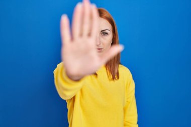 Young woman standing over blue background doing stop sing with palm of the hand. warning expression with negative and serious gesture on the face. 