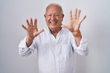 Gri saçlı yaşlı adam izole edilmiş arka planda duruyor ve parmaklarıyla on numarayı gösterirken kendinden emin ve mutlu bir şekilde gülümsüyor.. 