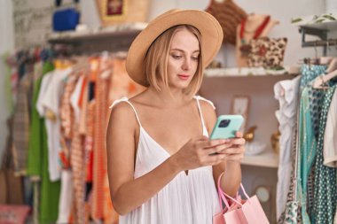 Young blonde woman customer wearing summer hat using smartphone at clothing store