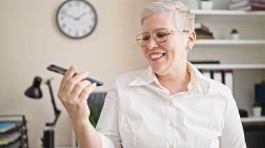 Orta yaşlı, kır saçlı, iş kadını. Ofiste çalışan akıllı telefondan sesli mesaj gönderiyor.