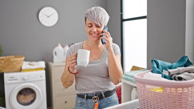 Akıllı telefondan konuşan, kahve içen çamaşırhanede gülümseyen genç bir kadın.