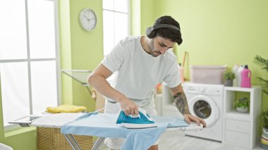 Genç İspanyol adam müzik dinliyor çamaşırhanede giysileri ütülüyor.