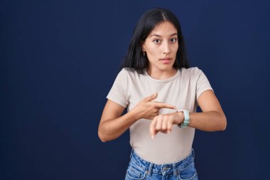 Young hispanic woman standing over blue background in hurry pointing to watch time, impatience, upset and angry for deadline delay 